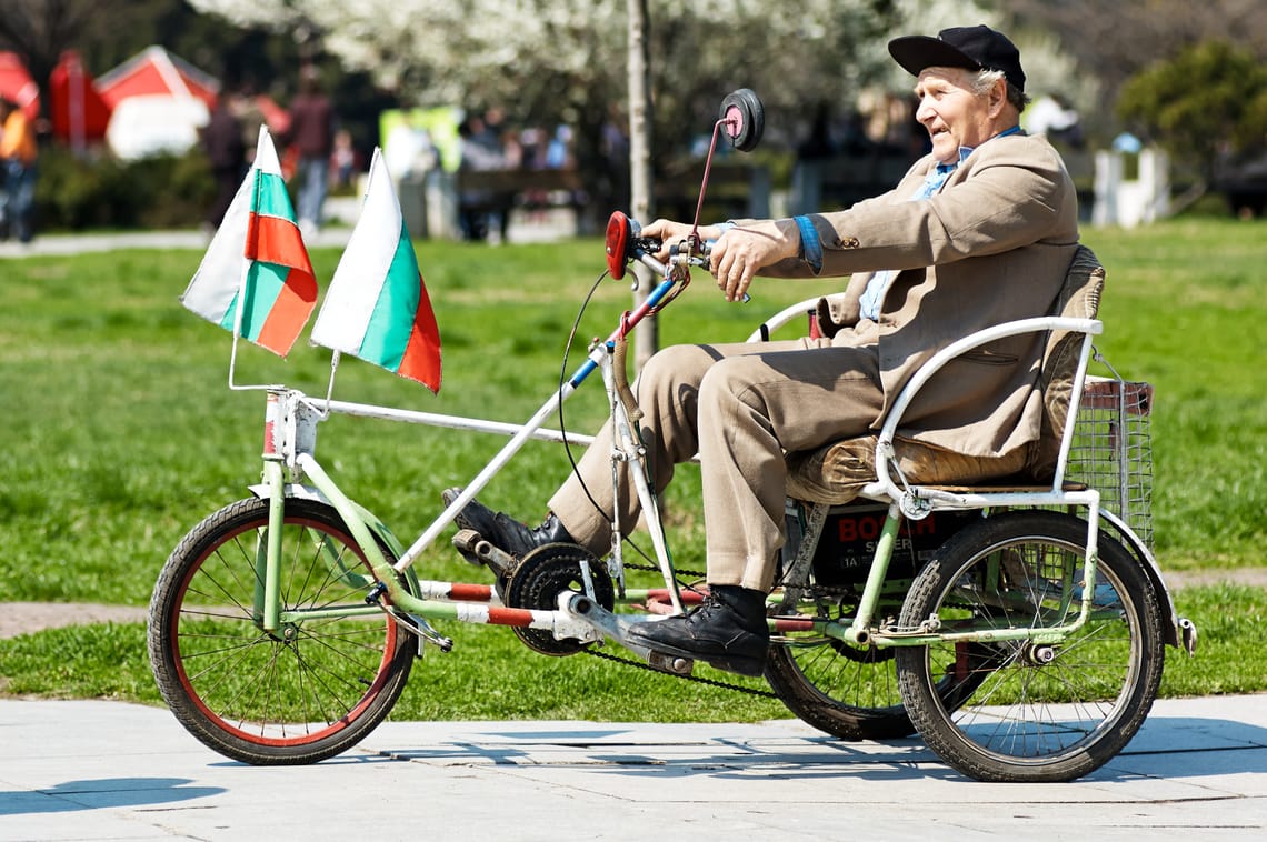 An elderly man wearing a cap and suit jacket riding a handmade three-wheeled recumbent bike with two small Bulgarian flags on the front in a sunny park.