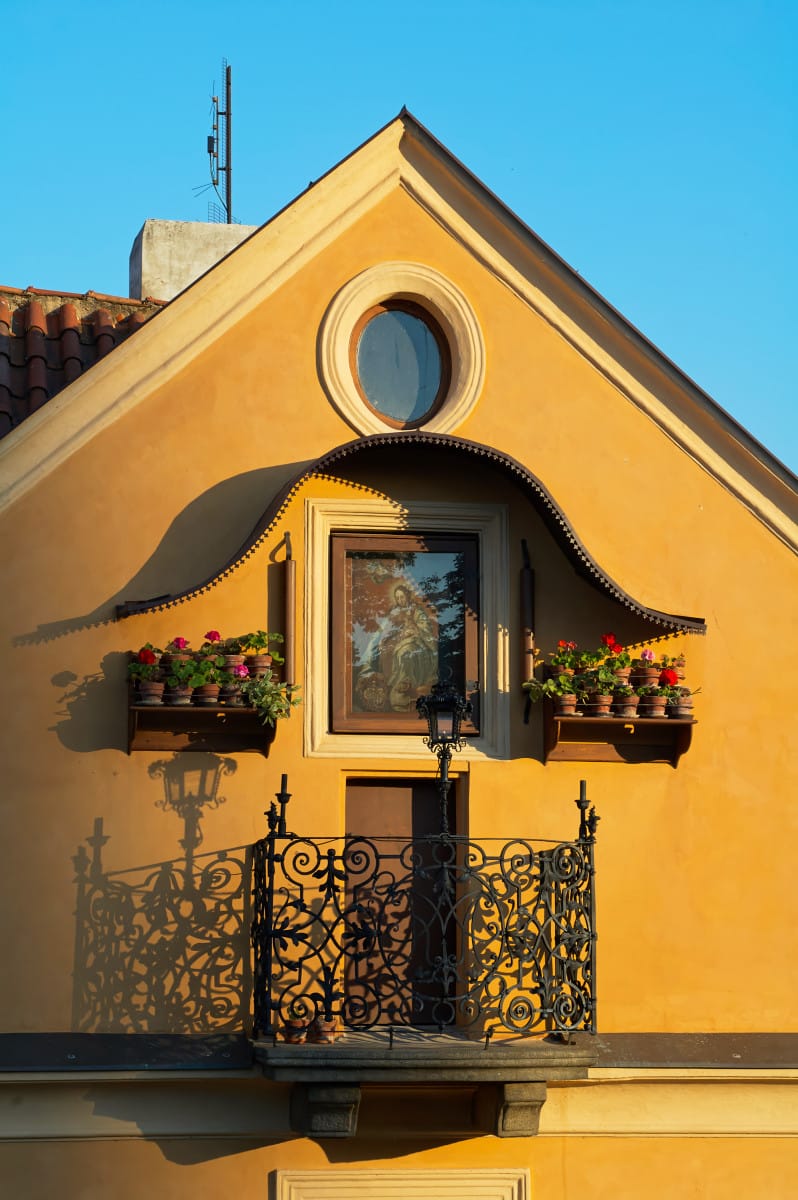 Yellow building façade with a wrought iron balcony, flower boxes, and a framed painting in warm sunlight.