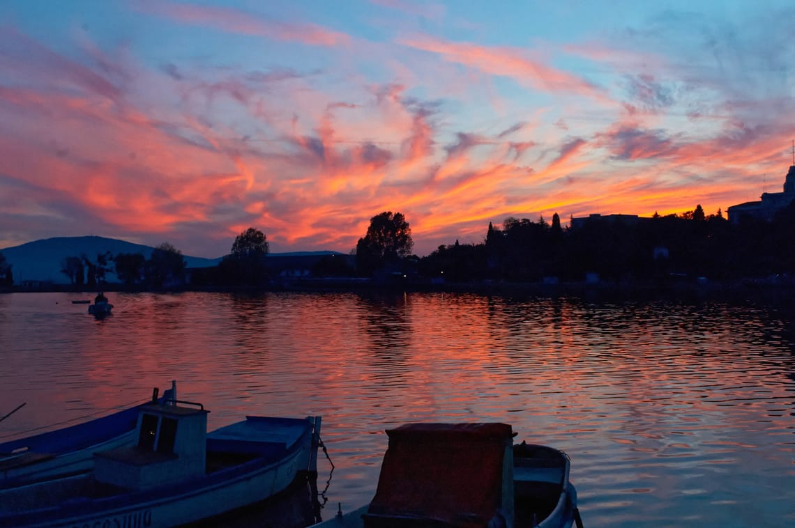 Small boats moored on calm water at sunset with vibrant pink and orange clouds reflected on the surface.
