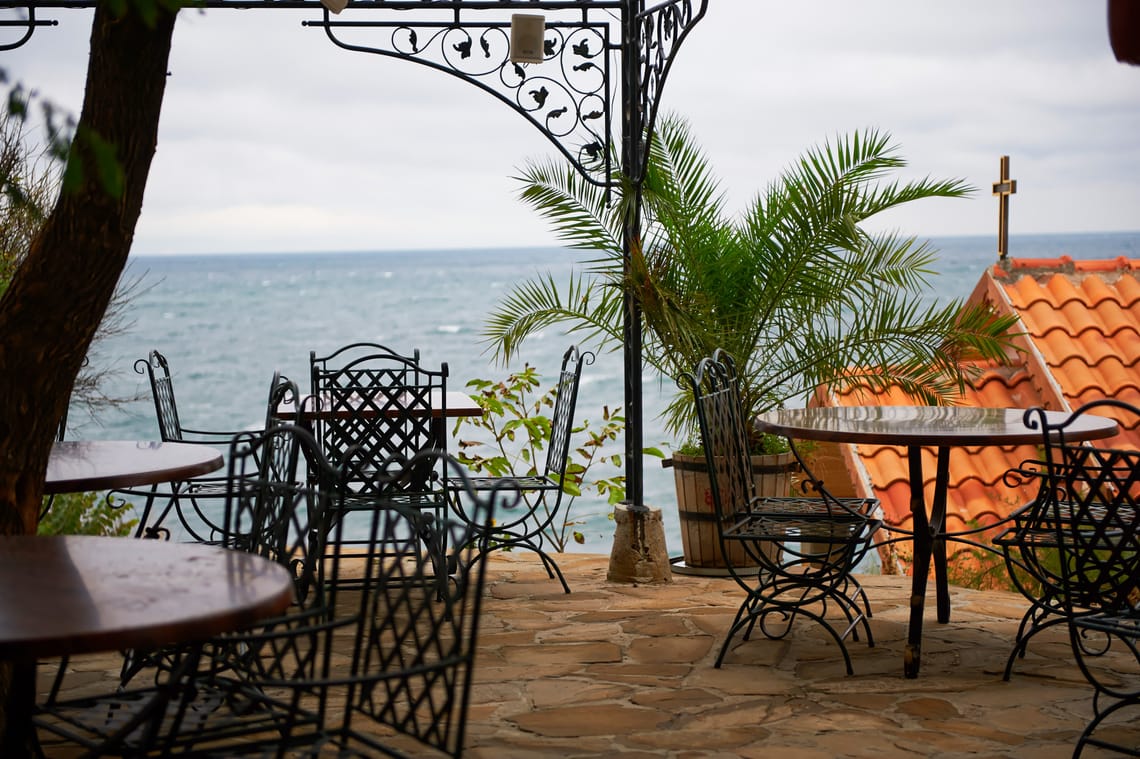 An atmospheric autumn view of Sozopol featuring an empty seaside terrace with wrought-iron furniture overlooking a stormy sea and a small chapel.
